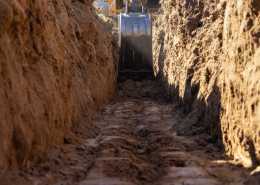 A yellow excavator operates in a deep trench with tall dirt walls on either side and muddy ground below.