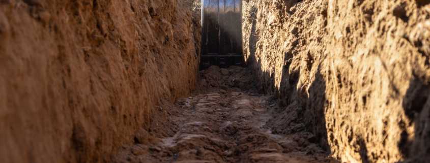 A yellow excavator operates in a deep trench with tall dirt walls on either side and muddy ground below.