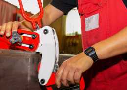 A worker wearing red overalls and a black t-shirt hooks an industrial lifting clamp to a heavy object.