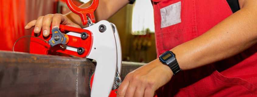 A worker wearing red overalls and a black t-shirt hooks an industrial lifting clamp to a heavy object.
