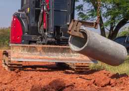A red excavator with a large blade, using a metal hook to lift a cylindrical concrete pipe outdoors over red dirt.