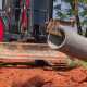 A red excavator with a large blade, using a metal hook to lift a cylindrical concrete pipe outdoors over red dirt.
