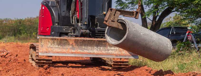A red excavator with a large blade, using a metal hook to lift a cylindrical concrete pipe outdoors over red dirt.