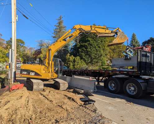 CAT excavator loading precast barriers onto truck using blue scissor clamp at side of roadway with blue sky