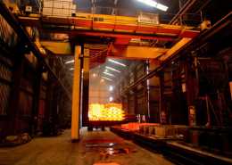 A large group of forged steel rods is outside a furnace in an industrial factory setting, with high ceilings and ample space.