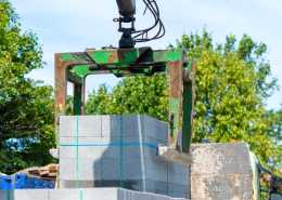A green mechanical grab lifts a pallet of concrete blocks at an outdoor construction site surrounded by green trees
