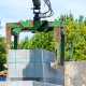 A green mechanical grab lifts a pallet of concrete blocks at an outdoor construction site surrounded by green trees