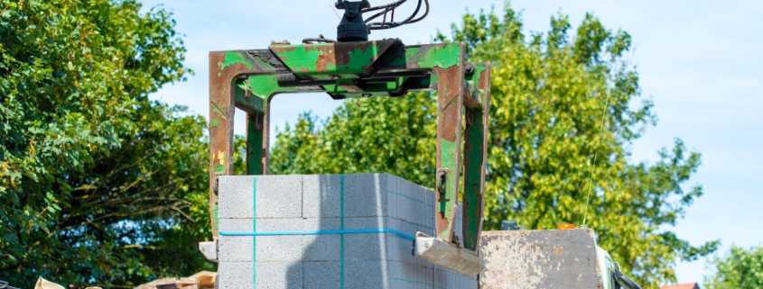 A green mechanical grab lifts a pallet of concrete blocks at an outdoor construction site surrounded by green trees
