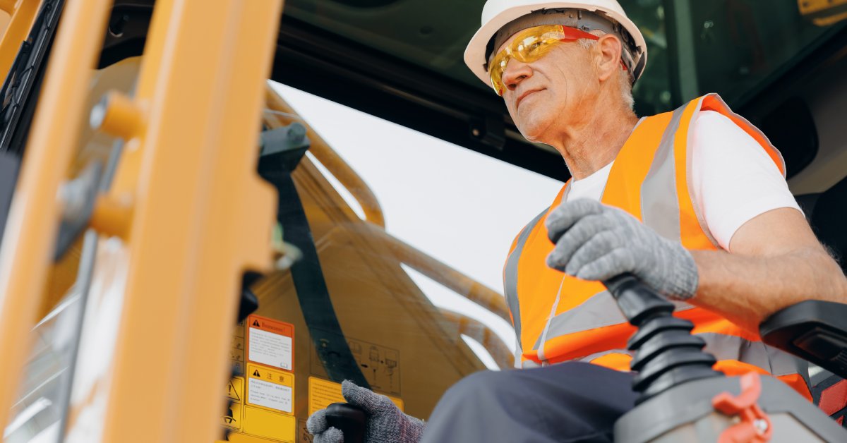 A smiling construction worker wearing safety gear and an orange vest controls an excavator machine on site.