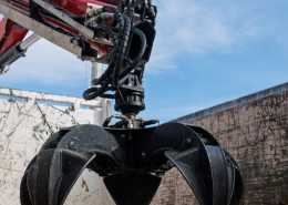 Heavy machinery with a black grapple claw attachment mounted on a red hydraulic arm against a clear blue sky.