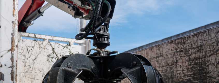 Heavy machinery with a black grapple claw attachment mounted on a red hydraulic arm against a clear blue sky.