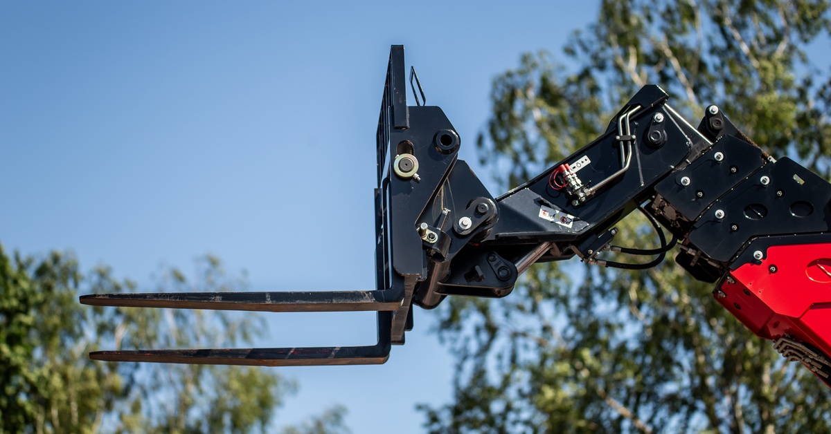 A black industrial forklift attachment on a hydraulic crane arm with metal prongs against a blue sky and green trees.