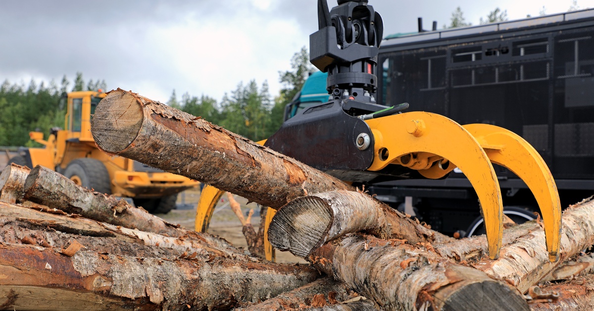 A large yellow and black grapple attachment lifts a log from a pile at a lumber yard with machinery in the background.