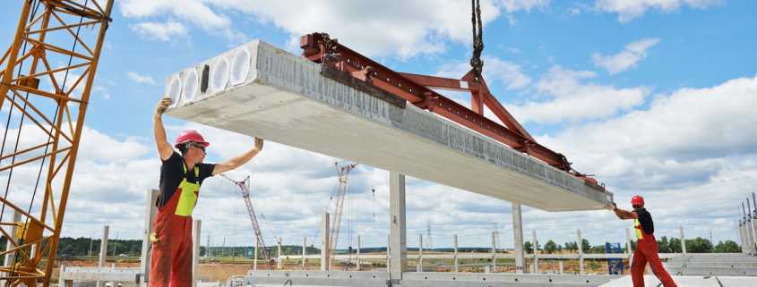 Two construction workers guide an industrial attachment lifting a concrete slab into place on a building site.