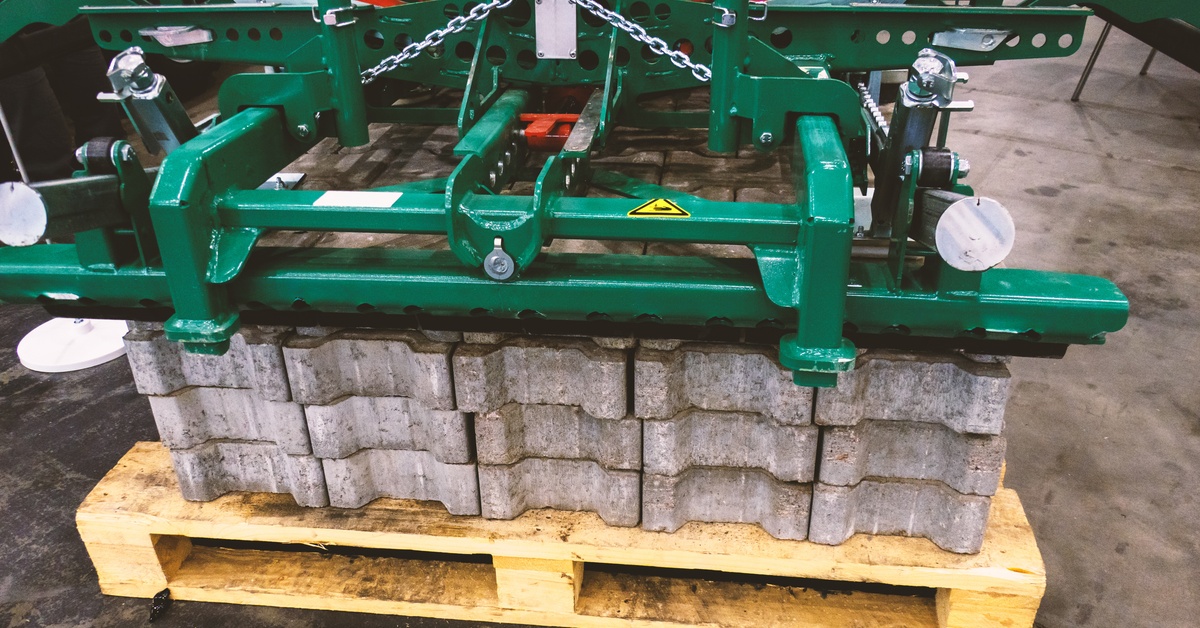 A green concrete block lifting device grips stacked pavers on a wooden pallet in a warehouse or workshop setting.