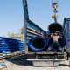 A worker guides a crane lifting large blue pipes onto a flatbed truck at a construction site with stacked pipes.