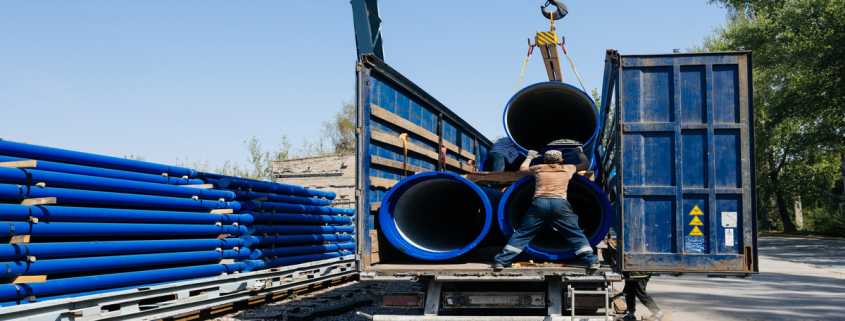 A worker guides a crane lifting large blue pipes onto a flatbed truck at a construction site with stacked pipes.