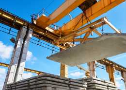 An overhead crane lifts a large concrete slab above stacked slabs at a construction site with blue skies above.