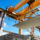 An overhead crane lifts a large concrete slab above stacked slabs at a construction site with blue skies above.