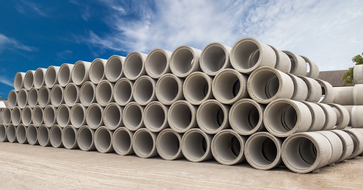 A large stack of precast concrete drainage pipes arranged in rows at an outdoor construction yard under a blue sky.
