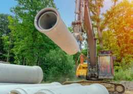 An excavator lifts a large precast concrete pipe at a construction site with stacked pipes and trees in the background.