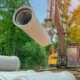 An excavator lifts a large precast concrete pipe at a construction site with stacked pipes and trees in the background.