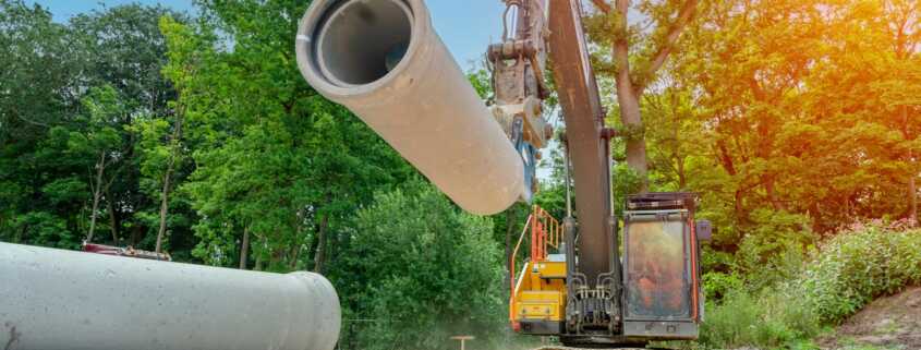 An excavator lifts a large precast concrete pipe at a construction site with stacked pipes and trees in the background.