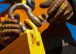 A construction worker secures a lifting shackle to a heavy-duty attachment point, wearing protective gloves and safety gear.