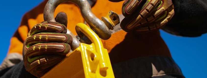 A construction worker secures a lifting shackle to a heavy-duty attachment point, wearing protective gloves and safety gear.