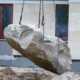 Large boulder suspended by lifting chains at a construction site with a tiled building in the background.