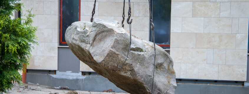 Large boulder suspended by lifting chains at a construction site with a tiled building in the background.