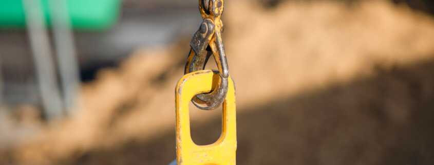 Yellow concrete block lifting hook connected to a heavy-duty steel chain above a precast barrier on an active job site.