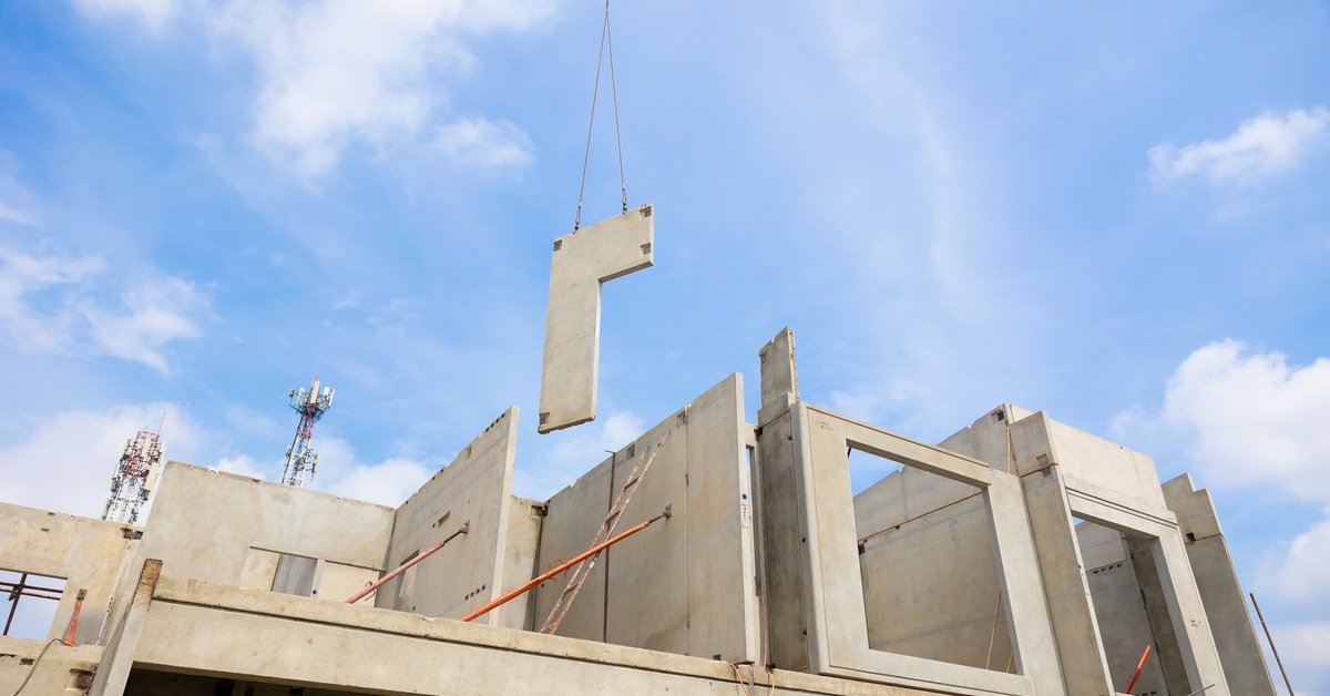 A crane lifts a precast concrete panel at an active construction site with a partially built concrete structure.