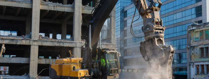 A yellow excavator runs a hydraulic shear attachment on a concrete demolition site surrounded by urban buildings.